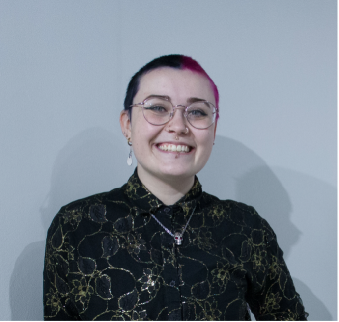 A square shot of Carly, a non-binary, white person with a buzzed haircut that is dyed half black and half pink. They are wearing a black button-up shirt with a gold floral pattern. They have round glasses and are smiling big. They have various facial piercings.