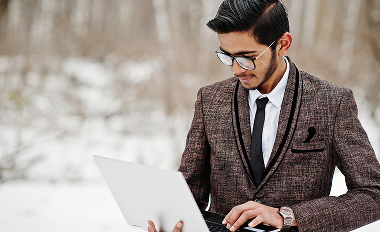 Indian student in suit holding laptop with snowing background.