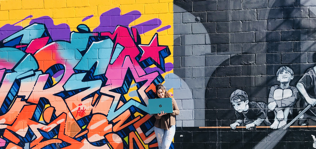 Summer Session 2020 Girl standing in front of murals outside holding a laptop