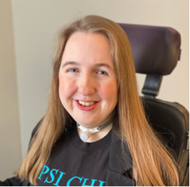 Smiling, Kaci, a 23-year-old white woman, has long, light brown hair and is wearing a black blazer and top, has a tracheostomy, and is seated in her power wheelchair.