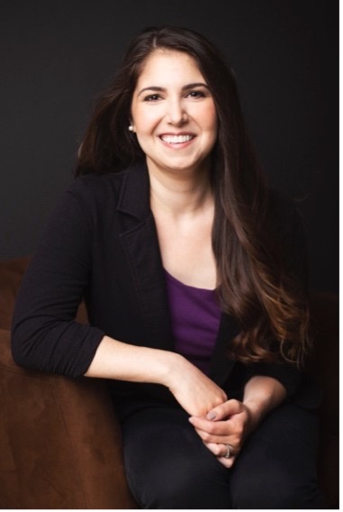 Marlee, a white cis woman, is smiling, has long dark brown hair. She is wearing a dark purple top, black blazer and black pants. She's seated on a brown chair, her hands are clasped.