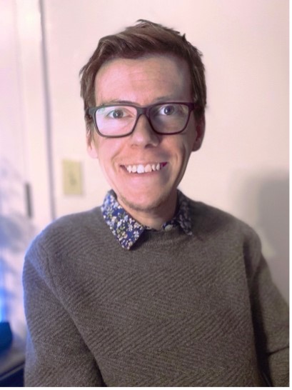 A photo from the mid-chest and up of Max, a white man in his 20s with dark grey squarish glasses, medium-short dark brown hair, and a short reddish-brown beard on his chin. He is wearing a grey sweater, and the collar of a dark blue shirt with a floral pattern is folded down over the neckline. He is smiling and sitting in front of a plain white wall that is slightly blurred in the background.