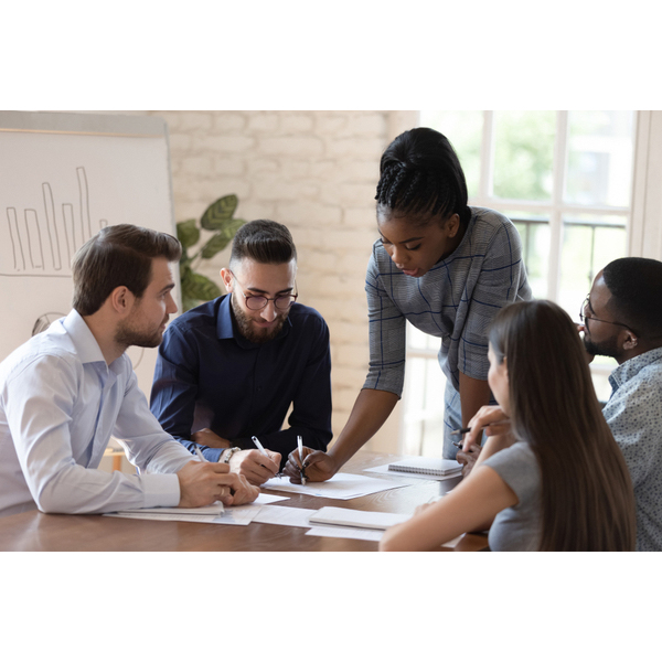 Woman stands at table in office reviewing paperwork while 3 men and 1 women sit and listen