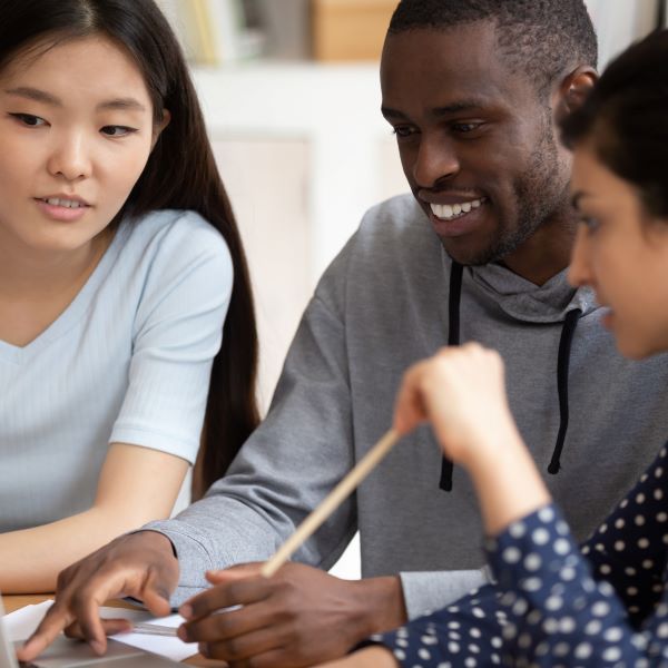 Two female Asian students and one male African-American student smile and sit together while facing a laptop screen.