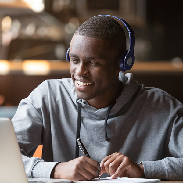 Smiling young male African student wearing headphones looking at laptop screen and writing notes in cafe