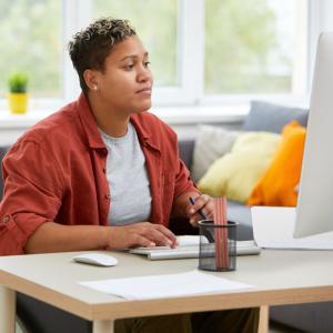 A woman in a wheelchair sits at her desktop computer. She wears an orange shirt over a white tee shirt.