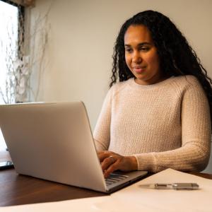 Woman with long dark curly hair and white shirt typing on a white laptop.