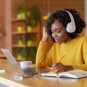 Black woman with short curly hair sits in front of a laptop wearing white headphones and writing in a blue notebook.