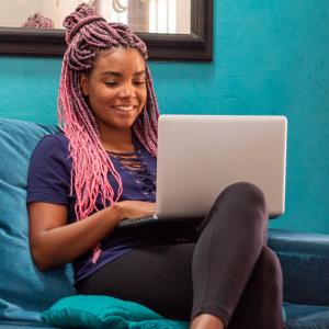 A woman seated on a teal colored couch, focused on her laptop. She has long pink hair.