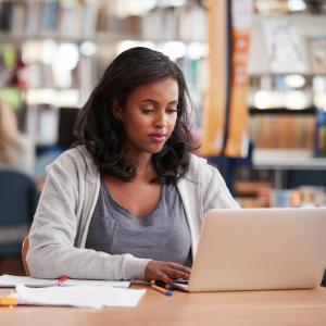 A woman with dark hair is seated in a library working on a laptop with papers and pens next to her.