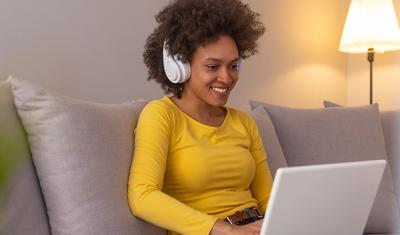 Young woman on couch using headphones and a laptop.