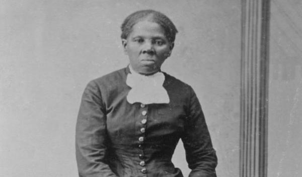 Photograph shows a full-length portrait of Harriet Tubman (1820?-1913) looking directly at the camera with folded hands resting on back of an upholstered chair.