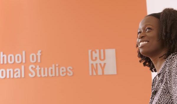 CUNY SPS Student Holding Laptop Smiles with School Sign Behind Her