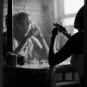 A brown skinned woman sits in front of a mirror near a window, with arms on a table and cigarette smoke in the air.