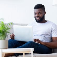 A man seated on a white couch smiles at his laptop computer. A green plant is in the foreground.