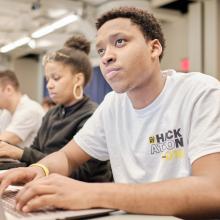 Young black man with a laptop wearing a CUNY Hackathon 2018 t-shirt