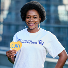 Student wearing CUNY SPS t-shirt and smiling while holding CUNY heart cut-out