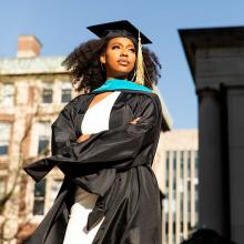 Female African American CUNY SPS graduate posing with arms folded and wearing cap and gown