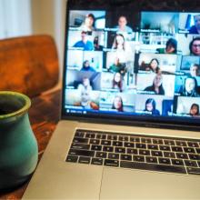 A video conference shows on a  laptop's screen. The laptop is on a desk with a coffee cup.