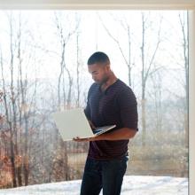 Man Using Laptop Computer While Standing Inside Against Glass Window With Snowy Scenery Outside